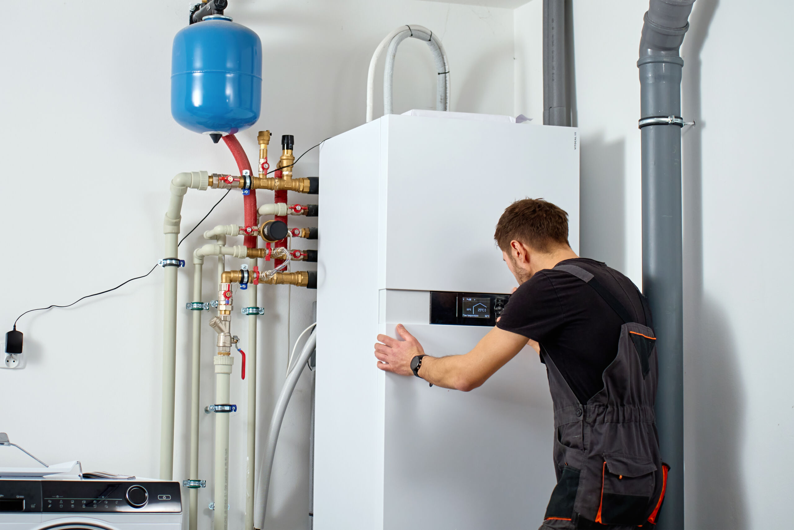 Technician installing indoor heat pump system with pipes and expansion tank. Man adjusting control panel on heat pump. Concept of home heating installation, energy efficiency and HVAC maintenance